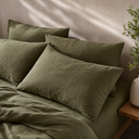 Four moss green linen pillowcases arranged on matching sheets in sunlit bedroom setting