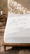 White linen fitted sheet on wooden bed frame with leafy branch shadows in sunlit bedroom